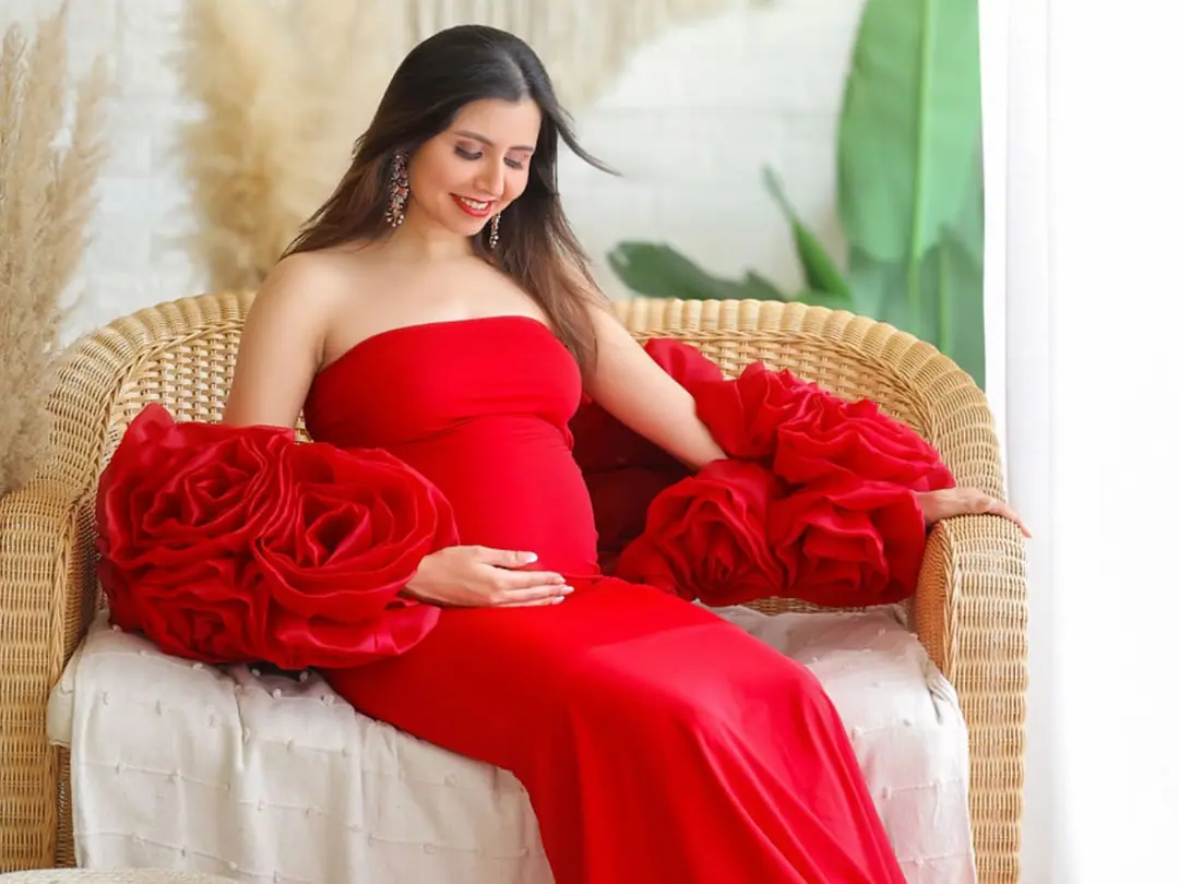 A smiling mother-to-be in a red gown on a wicker seat, holding her bump