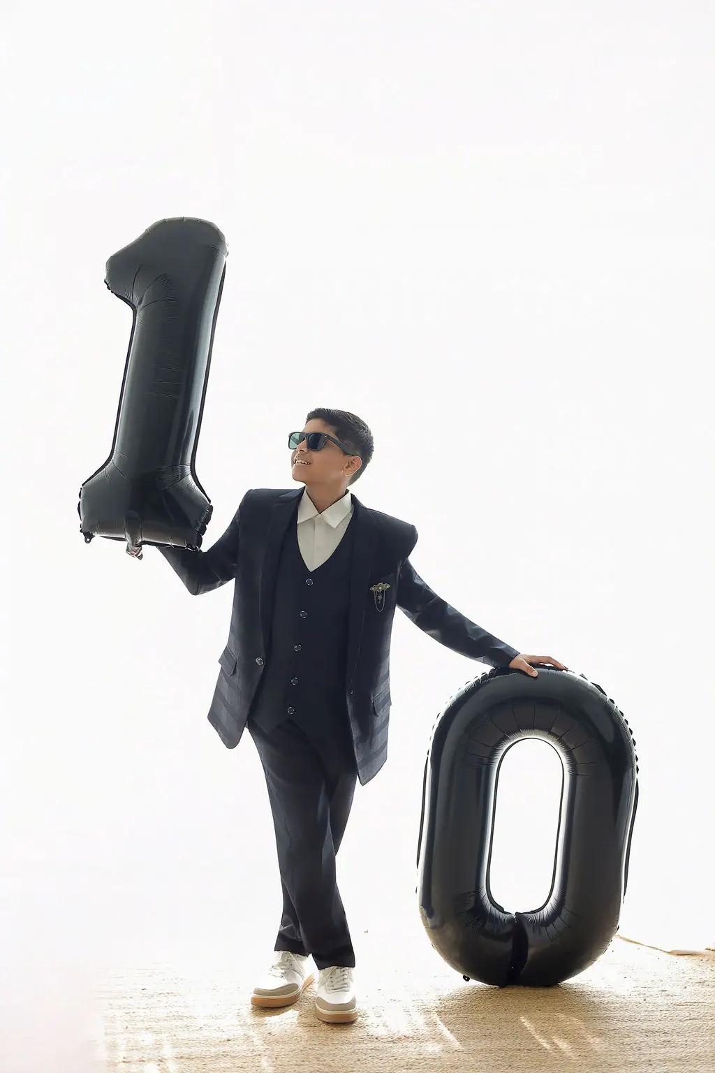 Child in a navy suit and sunglasses posing with giant black number balloons on a bright white set, birthday portrait at WonderBump Studio, Bengaluru