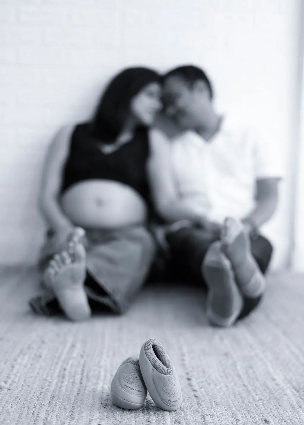 Tiny baby shoes in focus before a couple sitting close against a white wall, black and white couple maternity portrait at WonderBump Studio, Bengaluru