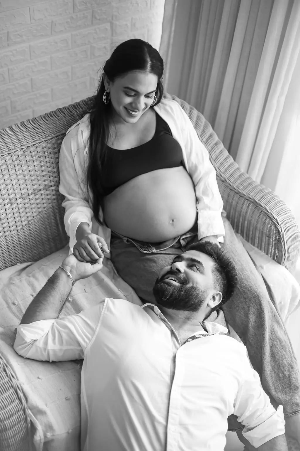 Expectant mother on a wicker chair holding hands with her partner resting in her lap, black and white couple maternity portrait at WonderBump Studio, Bengaluru