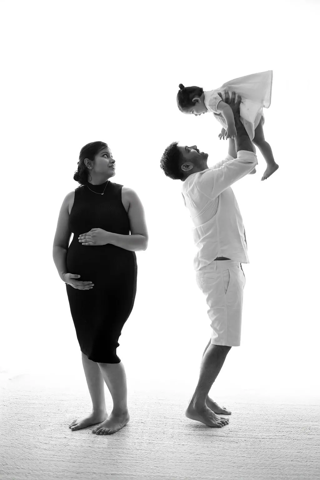 Expectant mother in a black dress watching the father lift their child high on a white set, black and white family portrait at WonderBump Studio, Bengaluru