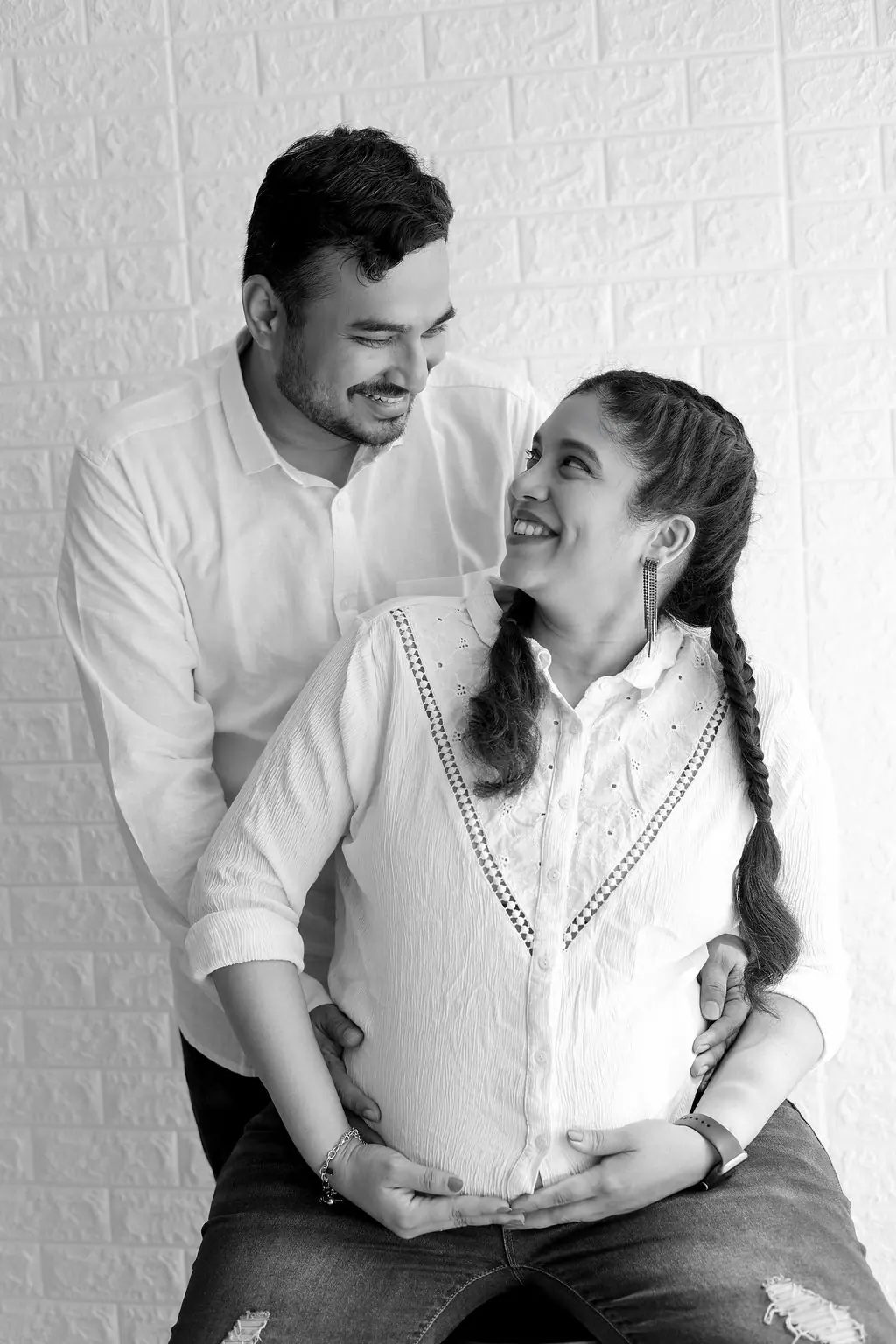Expectant mother with braids smiling up at the father behind her by a white brick wall, black and white maternity portrait at WonderBump Studio, Bengaluru