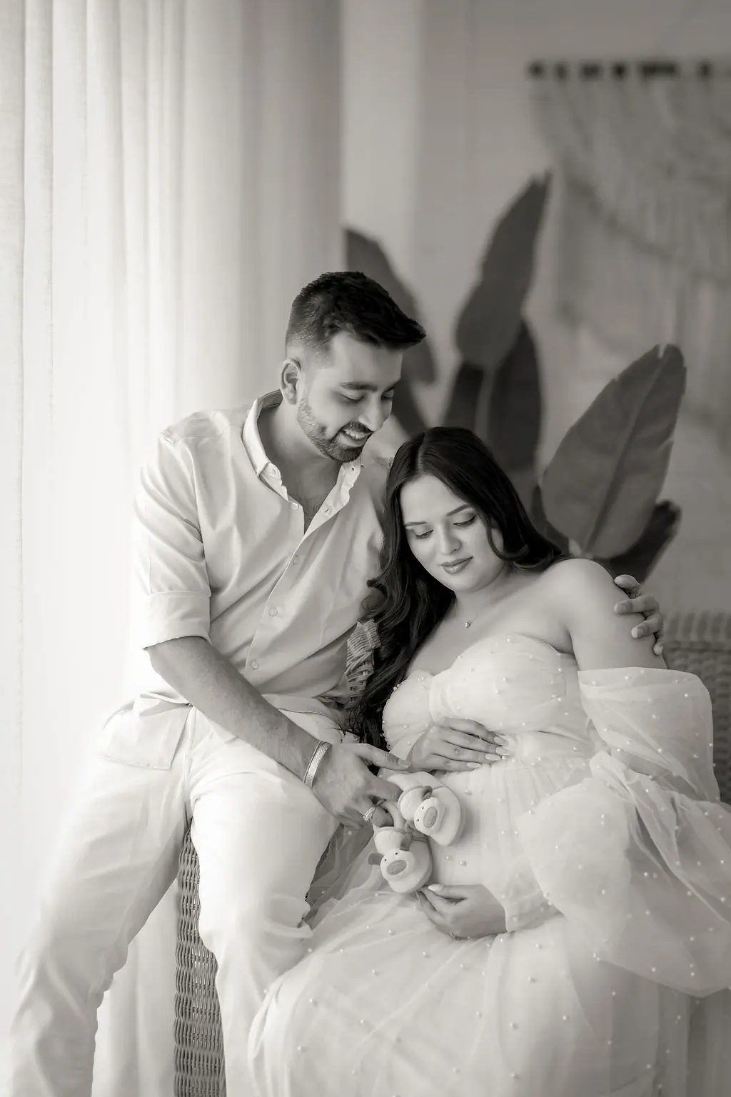 Father holding tiny baby shoes to the bump of an expectant mother in a tulle gown, black and white maternity portrait at WonderBump Studio, Bengaluru