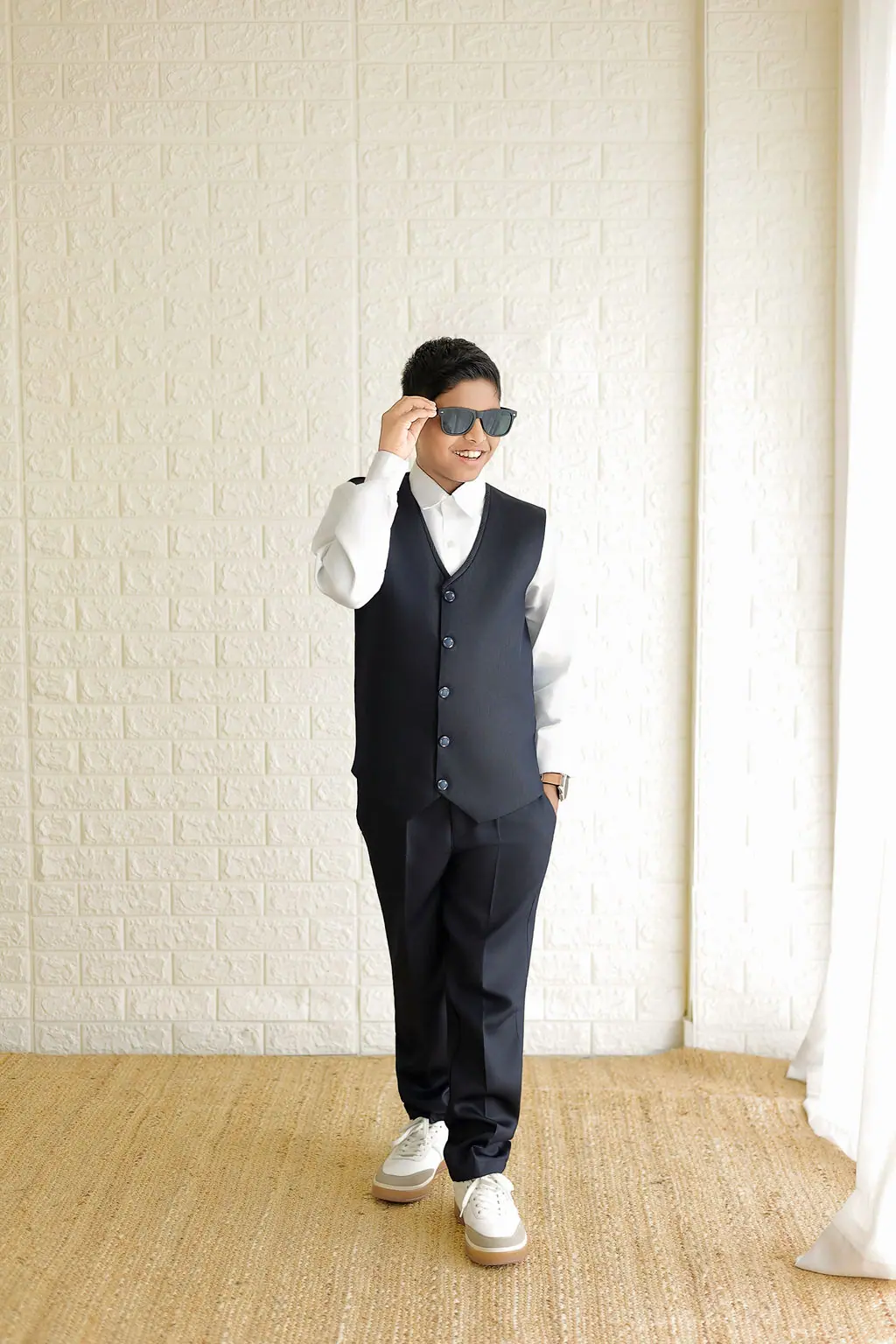 Smiling child in a navy waistcoat and white shirt adjusting sunglasses by a white brick wall, child portrait session at WonderBump Studio, Bengaluru