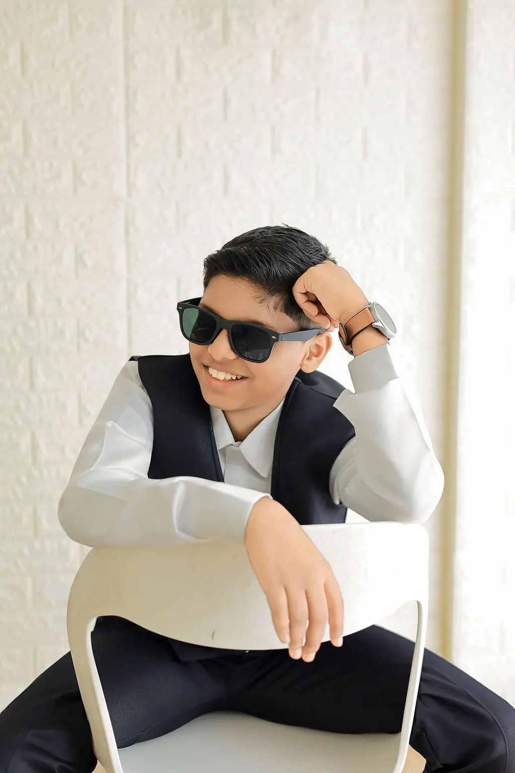 Grinning child in sunglasses and a navy waistcoat leaning on a white chair back, white brick wall, child portrait session at WonderBump Studio, Bengaluru