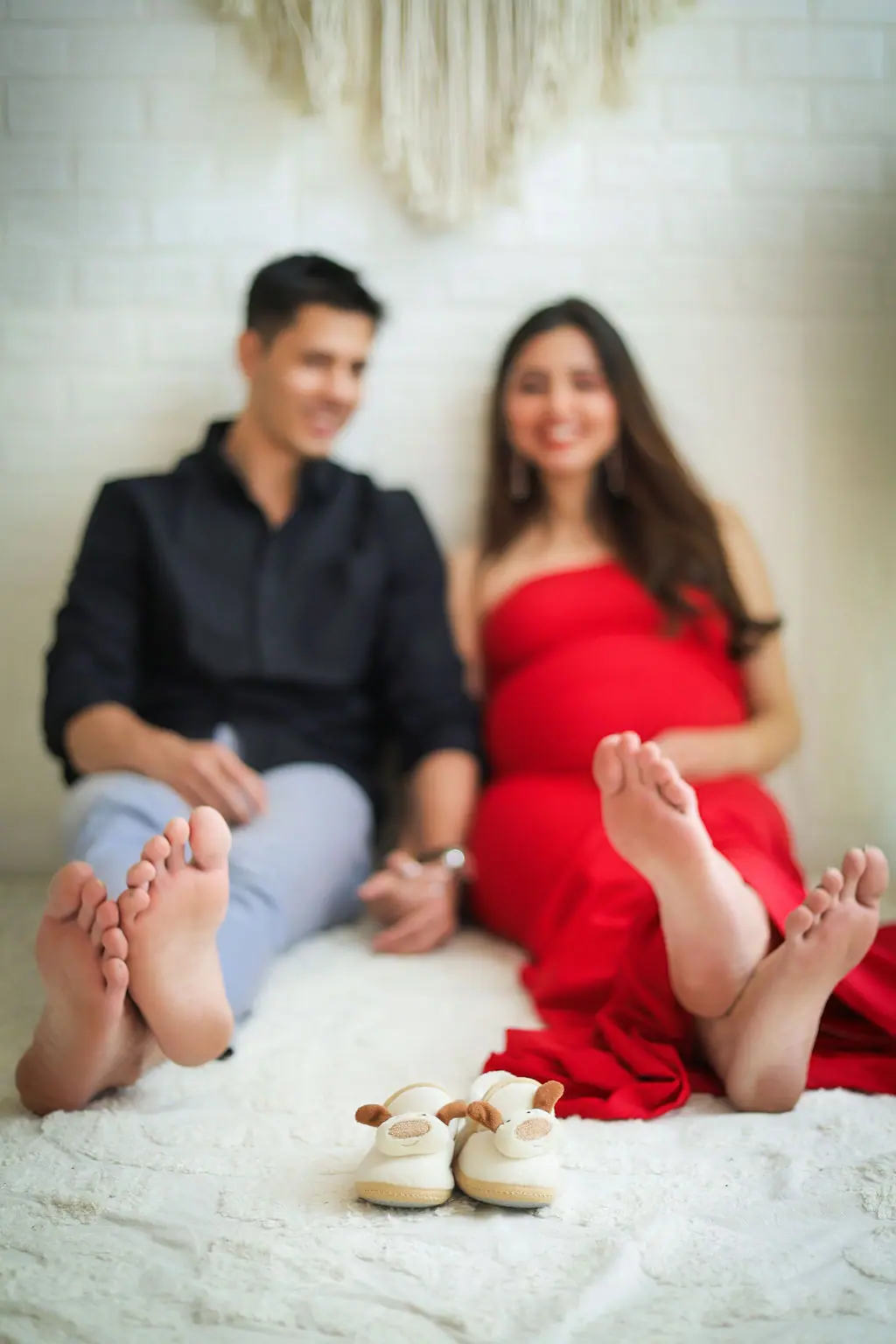 Tiny baby booties in focus before a barefoot couple, expectant mother in a red gown, couple maternity portrait at WonderBump Studio, Bengaluru