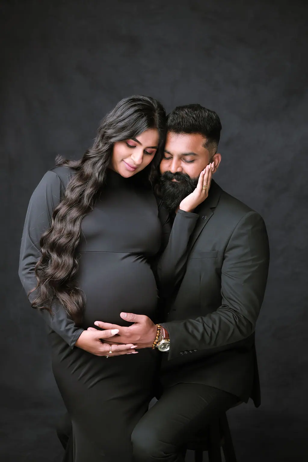 Expectant couple in black, cheek to cheek, cradling the bump on a dark textured backdrop, couple maternity portrait at WonderBump Studio, Bengaluru