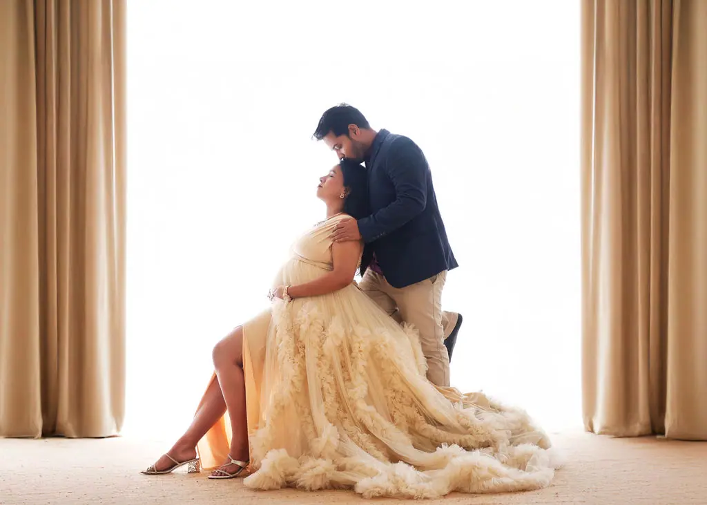 Expectant mother in a ruffled cream tulle gown receiving a forehead kiss between beige drapes, couple maternity portrait at WonderBump Studio, Bengaluru