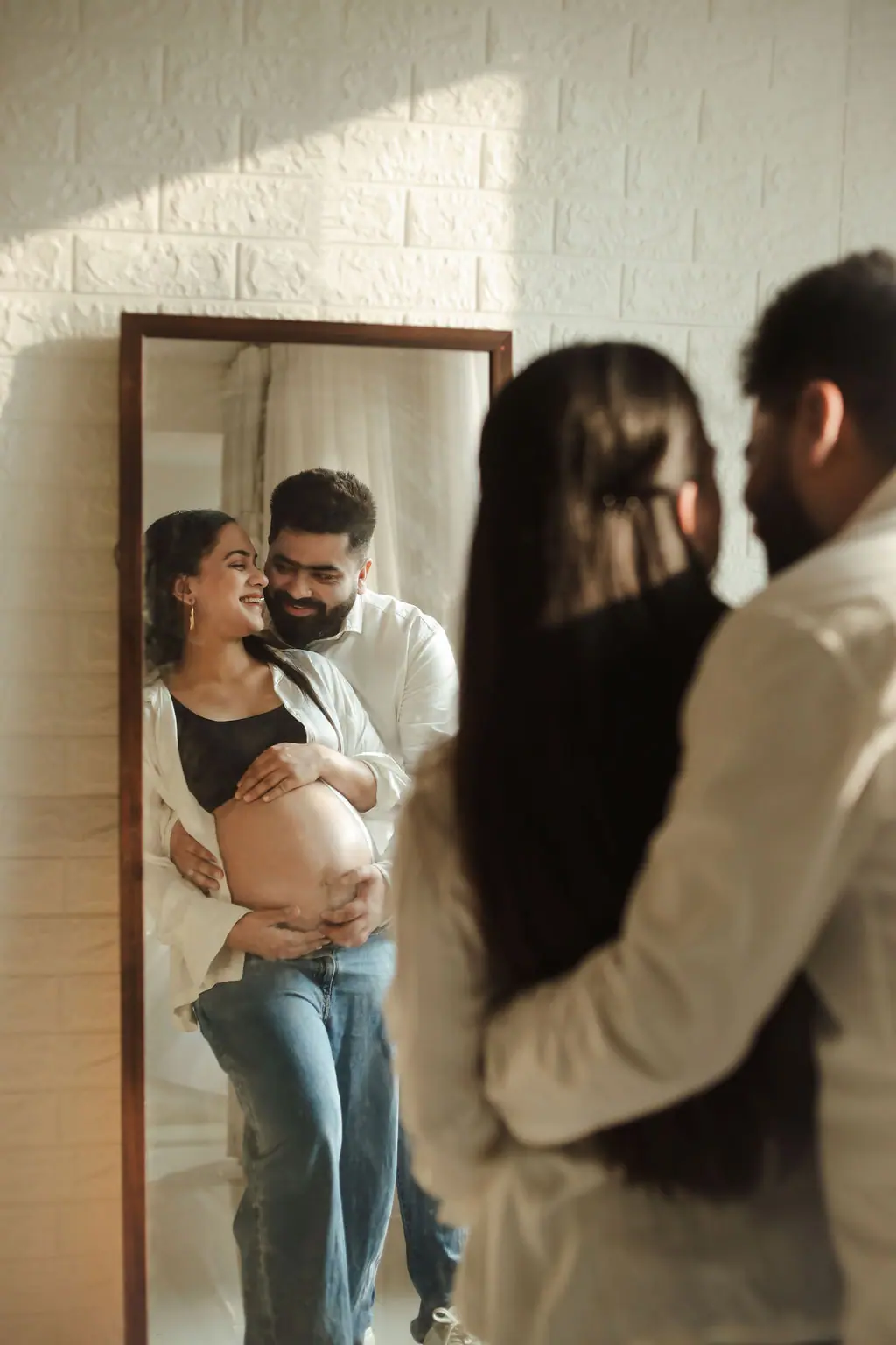 Expectant couple embracing before a tall mirror reflecting their smiles and bump, white brick wall, couple maternity portrait at WonderBump Studio, Bengaluru