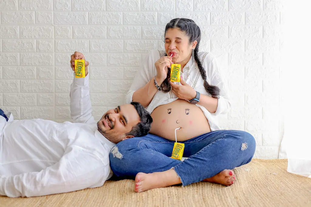 Father lying by the lap of an expectant mother with a face drawn on her bump, both with juice boxes, couple maternity portrait at WonderBump Studio, Bengaluru