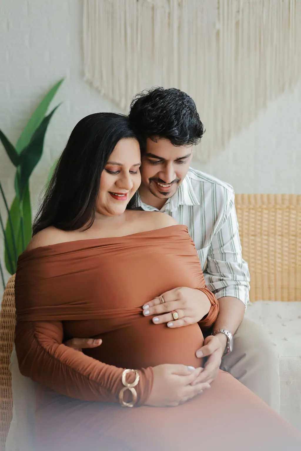 Father hugging an expectant mother in a rust off-shoulder gown on a rattan seat, couple maternity portrait at WonderBump Studio, Bengaluru