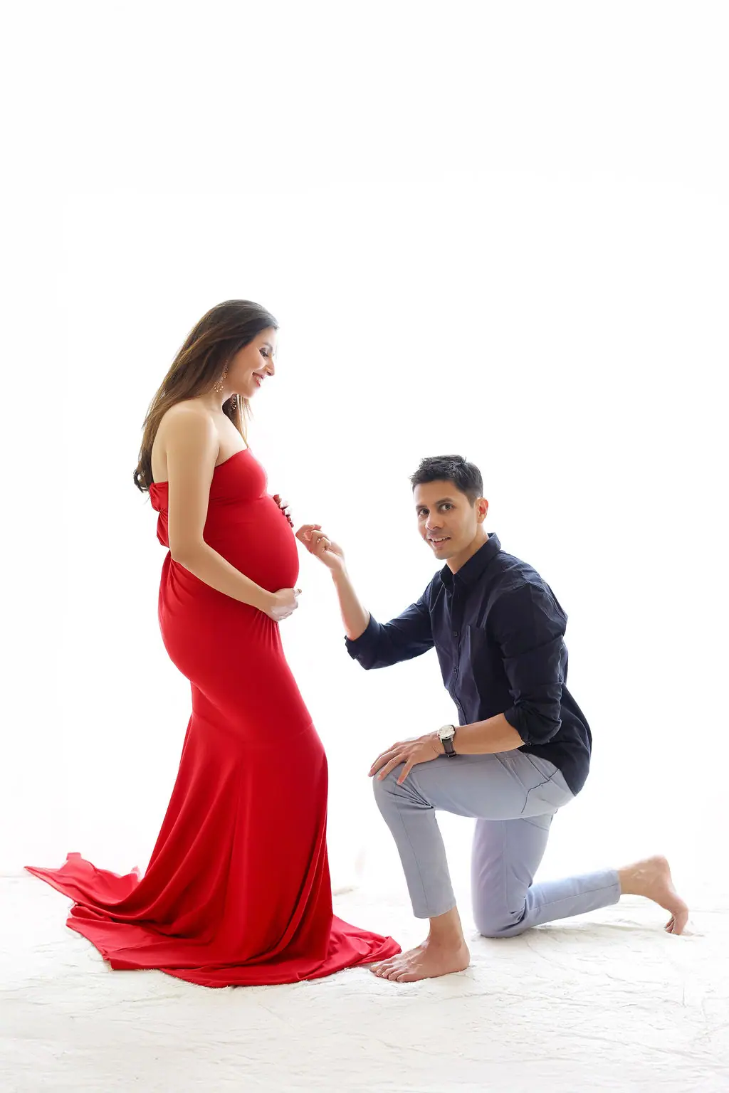 Father kneeling to hold hands with an expectant mother in a red strapless gown on white, couple maternity portrait at WonderBump Studio, Bengaluru