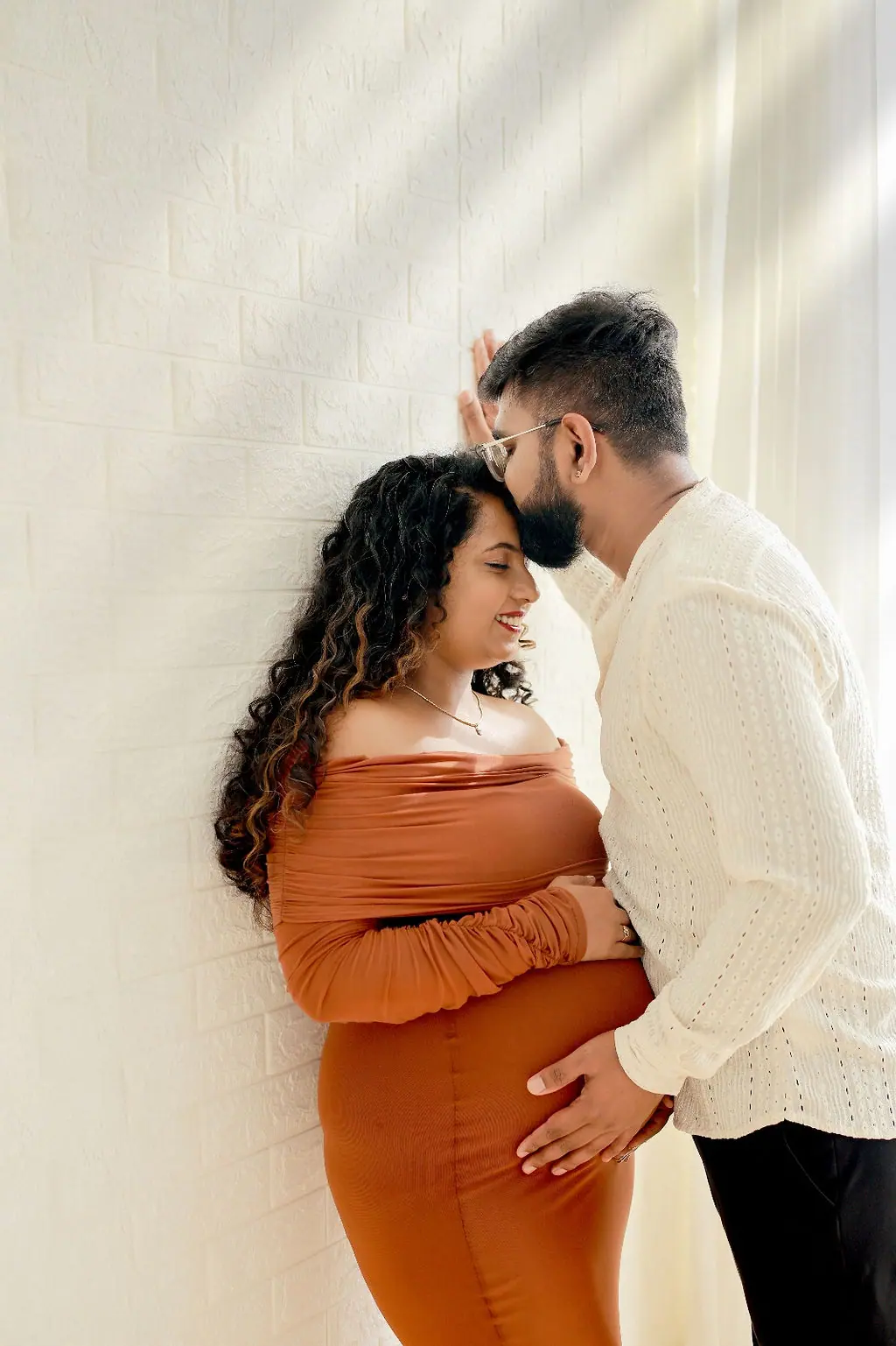 Father kissing the forehead of an expectant mother in a rust dress by a white brick wall, couple maternity portrait at WonderBump Studio, Bengaluru