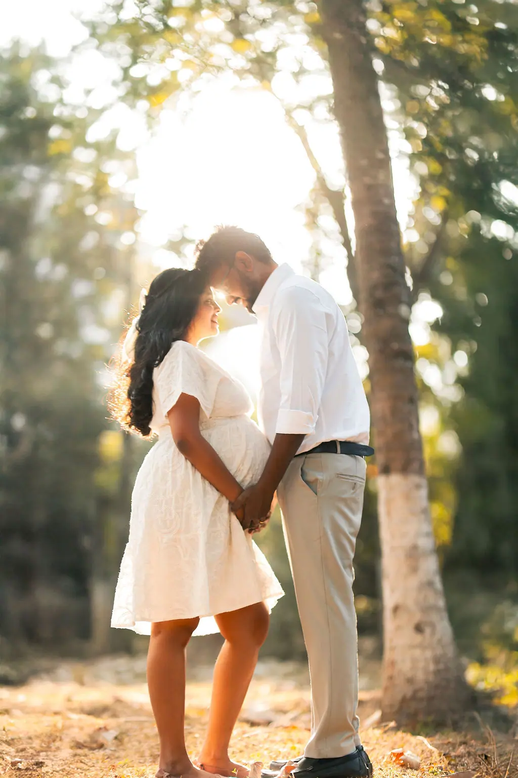 Expectant mother and partner touching foreheads under sunlit trees, white dress and shirt, couple maternity portrait by WonderBump Studio, Bengaluru