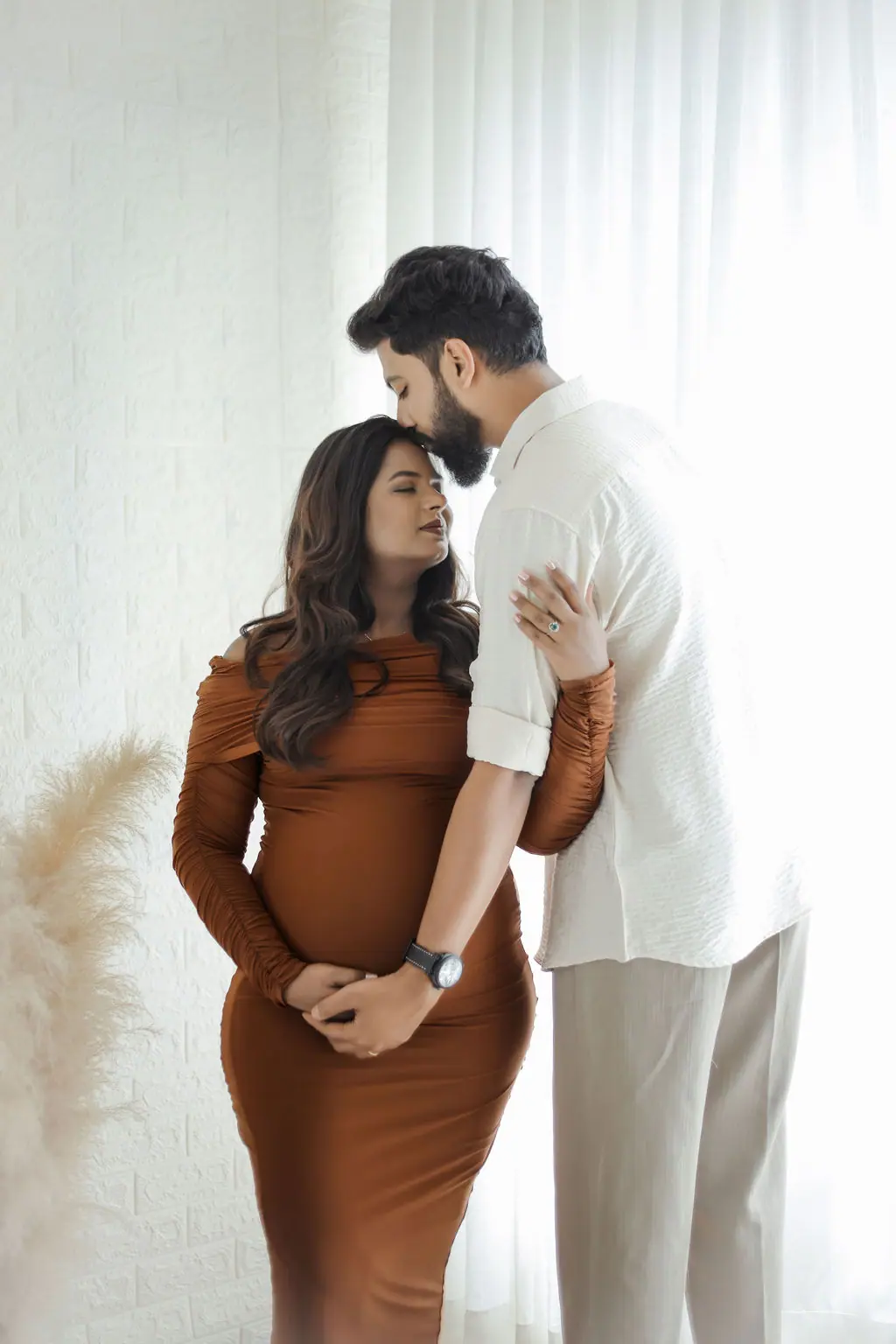 Expectant mother in a rust gown receiving a forehead kiss by white drapes and pampas grass, couple maternity portrait at WonderBump Studio, Bengaluru