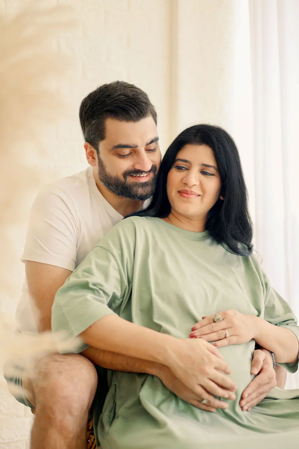 Expectant mother in a sage green tee cradling her bump as her partner hugs her from behind, couple maternity portrait at WonderBump Studio, Bengaluru