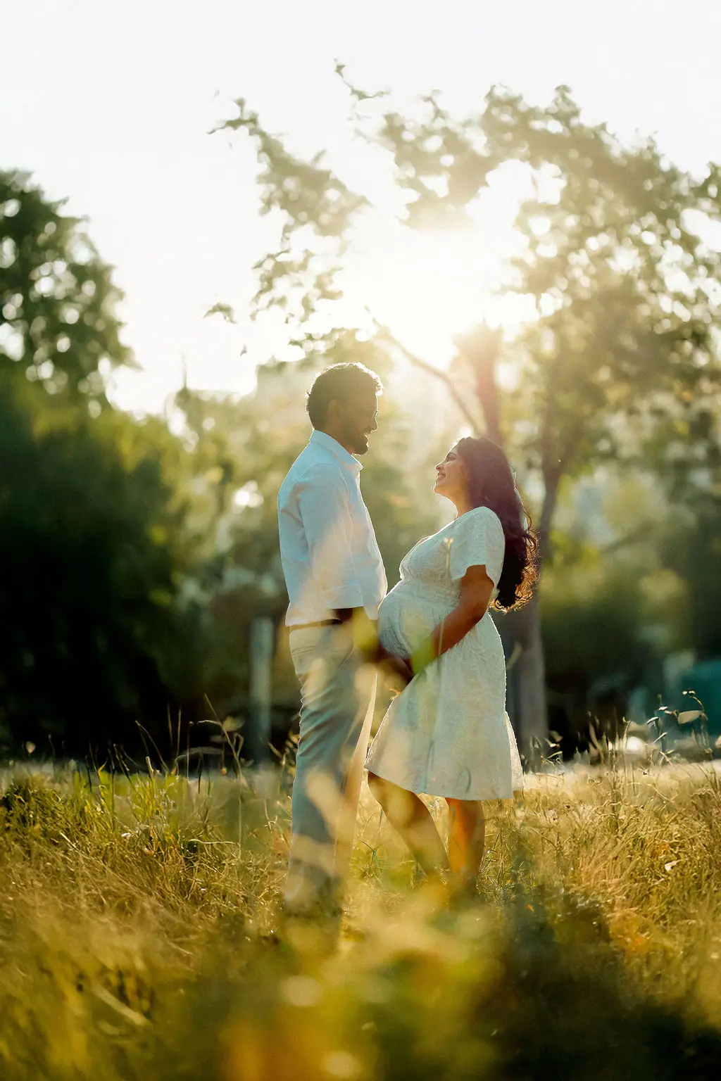 Expectant mother and partner smiling at each other in golden grass with sun behind, white outfits, couple maternity portrait by WonderBump Studio, Bengaluru