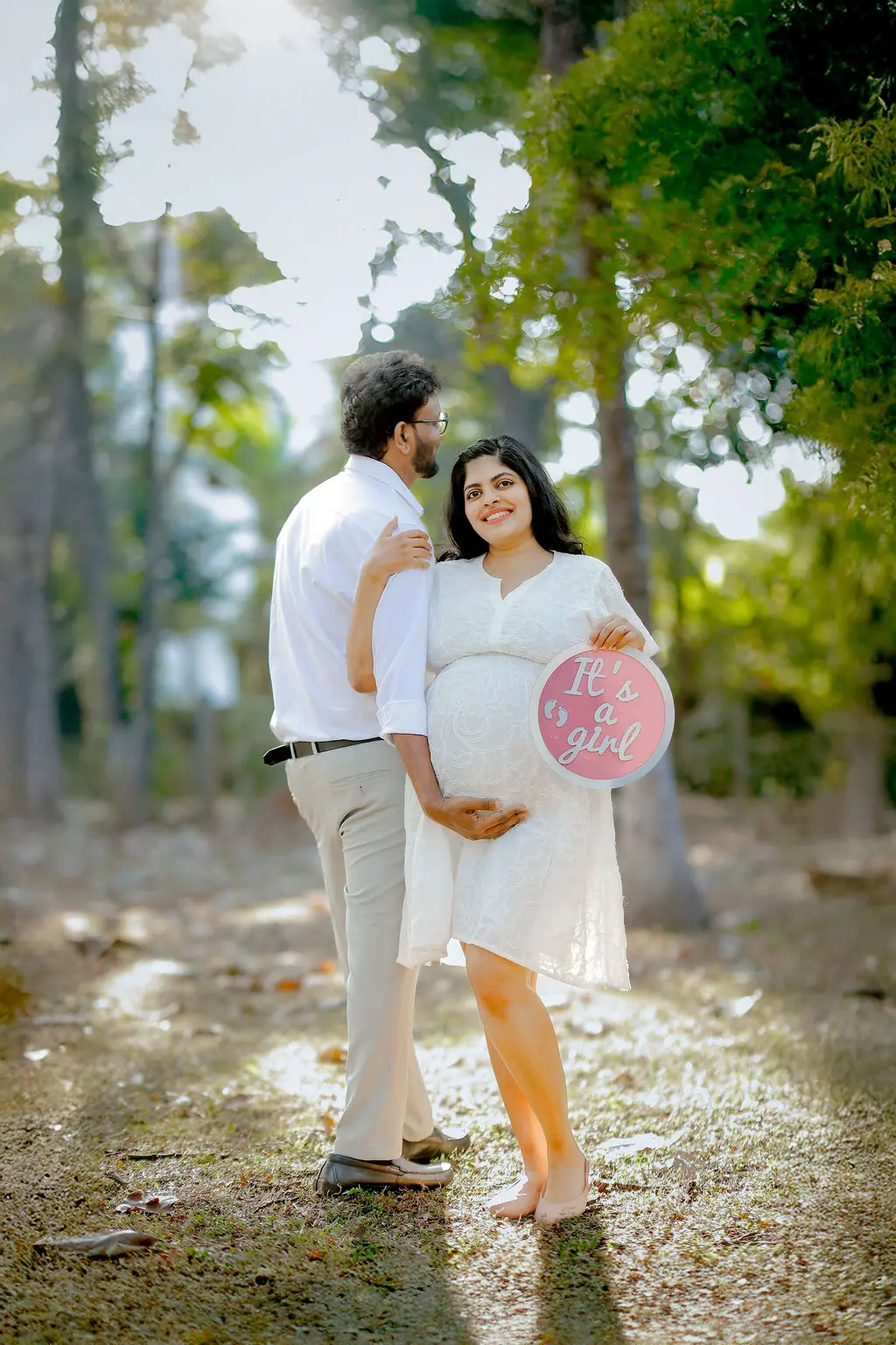 Smiling expectant mother with a pink baby announcement sign, partner's hand on her bump under trees, couple maternity portrait by WonderBump Studio, Bengaluru