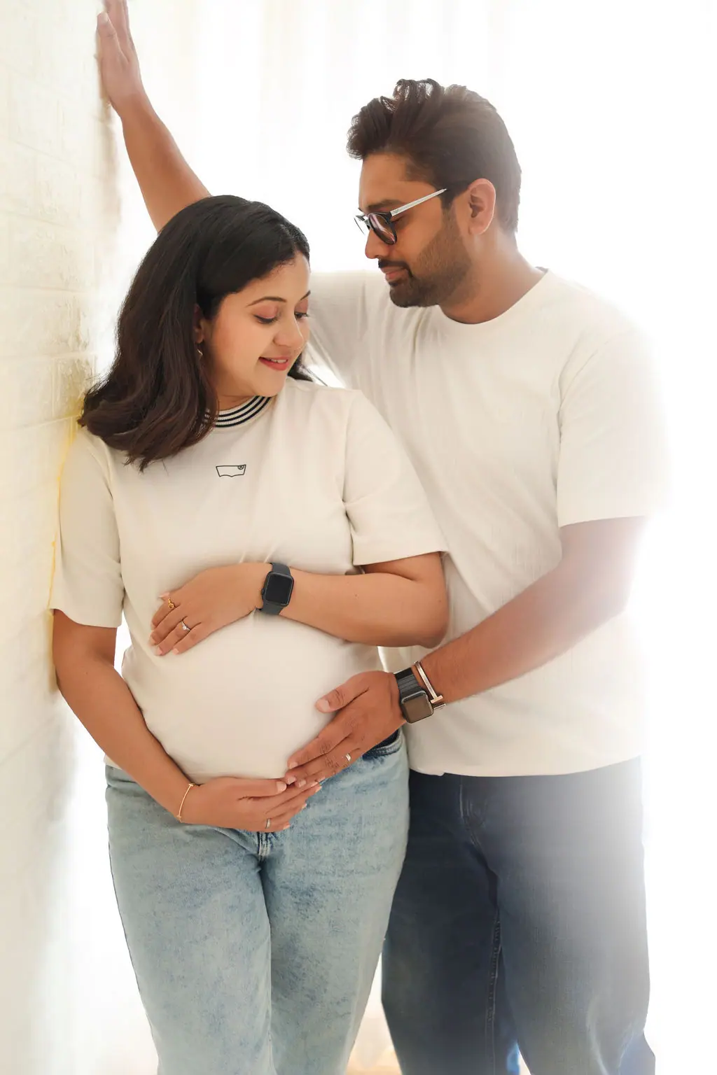 Expectant mother and partner in white tees and jeans cradling the bump against a white brick wall, couple maternity portrait at WonderBump Studio, Bengaluru