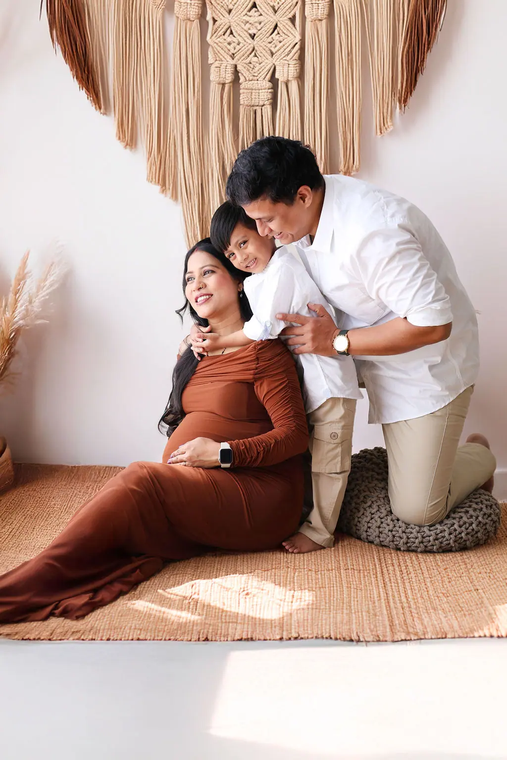 Expectant mother in a rust gown on a jute rug as the father and child hug her from behind, family maternity portrait at WonderBump Studio, Bengaluru