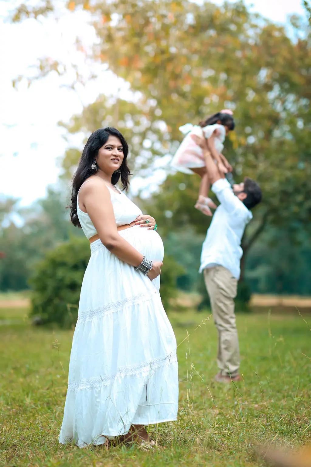 Expectant mother in a white tiered dress as the father lifts their child high on a grassy lawn, family maternity portrait by WonderBump Studio, Bengaluru
