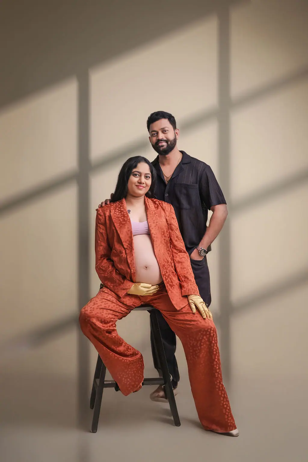 Expectant mother on a stool in a rust satin suit with the father behind her, window shadow backdrop, maternity portrait at WonderBump Studio, Bengaluru