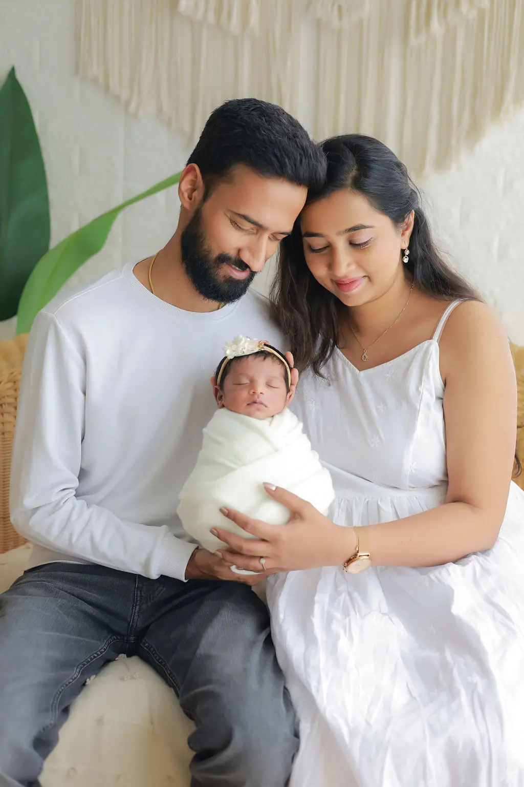 Parents with heads together holding their sleeping swaddled newborn in a floral headband, macramé wall, newborn family portrait at WonderBump Studio, Bengaluru