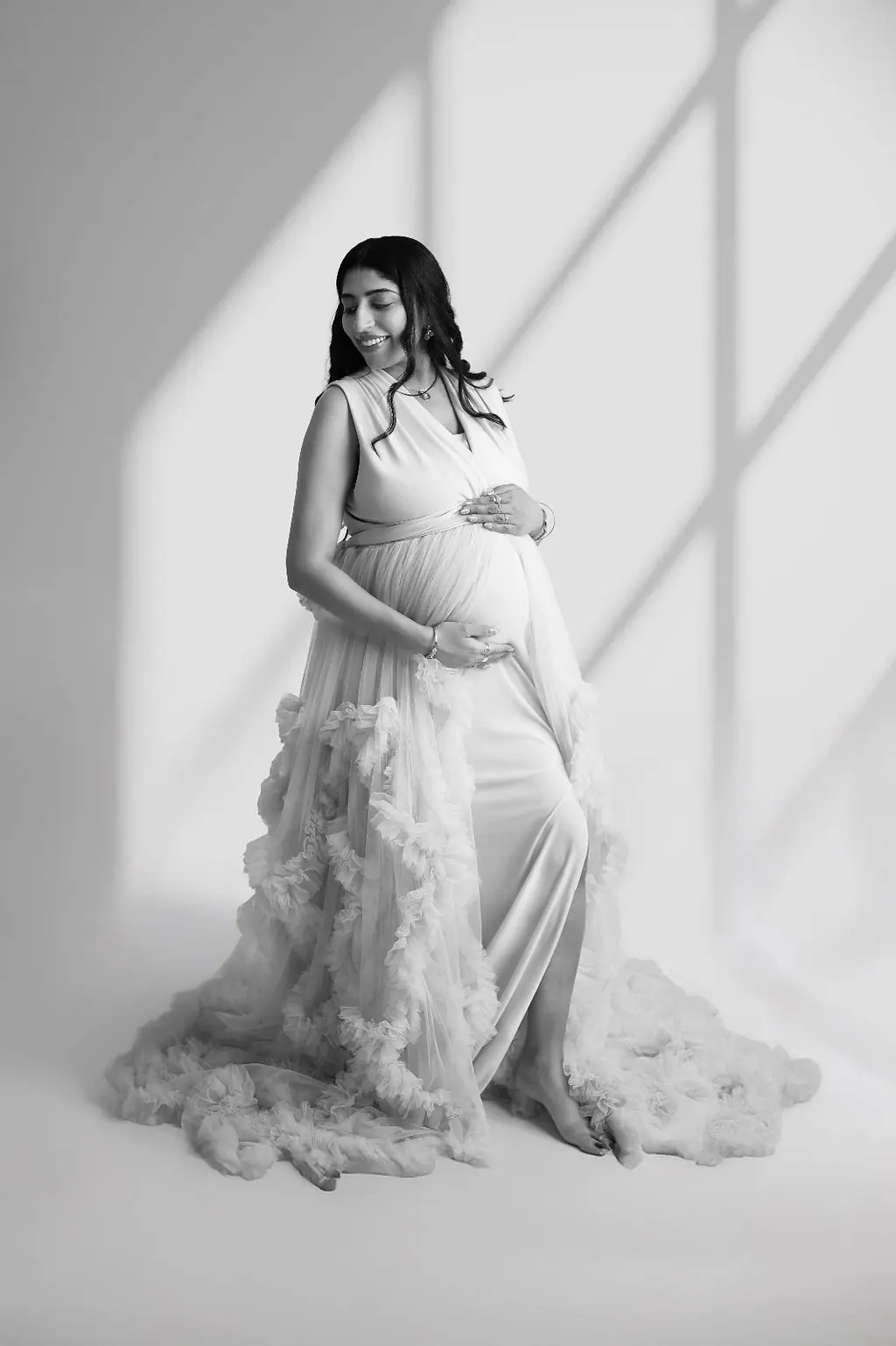 Expectant mother smiling at her bump in a ruffled tulle gown with window shadows, black and white maternity photoshoot at WonderBump Studio, Bengaluru