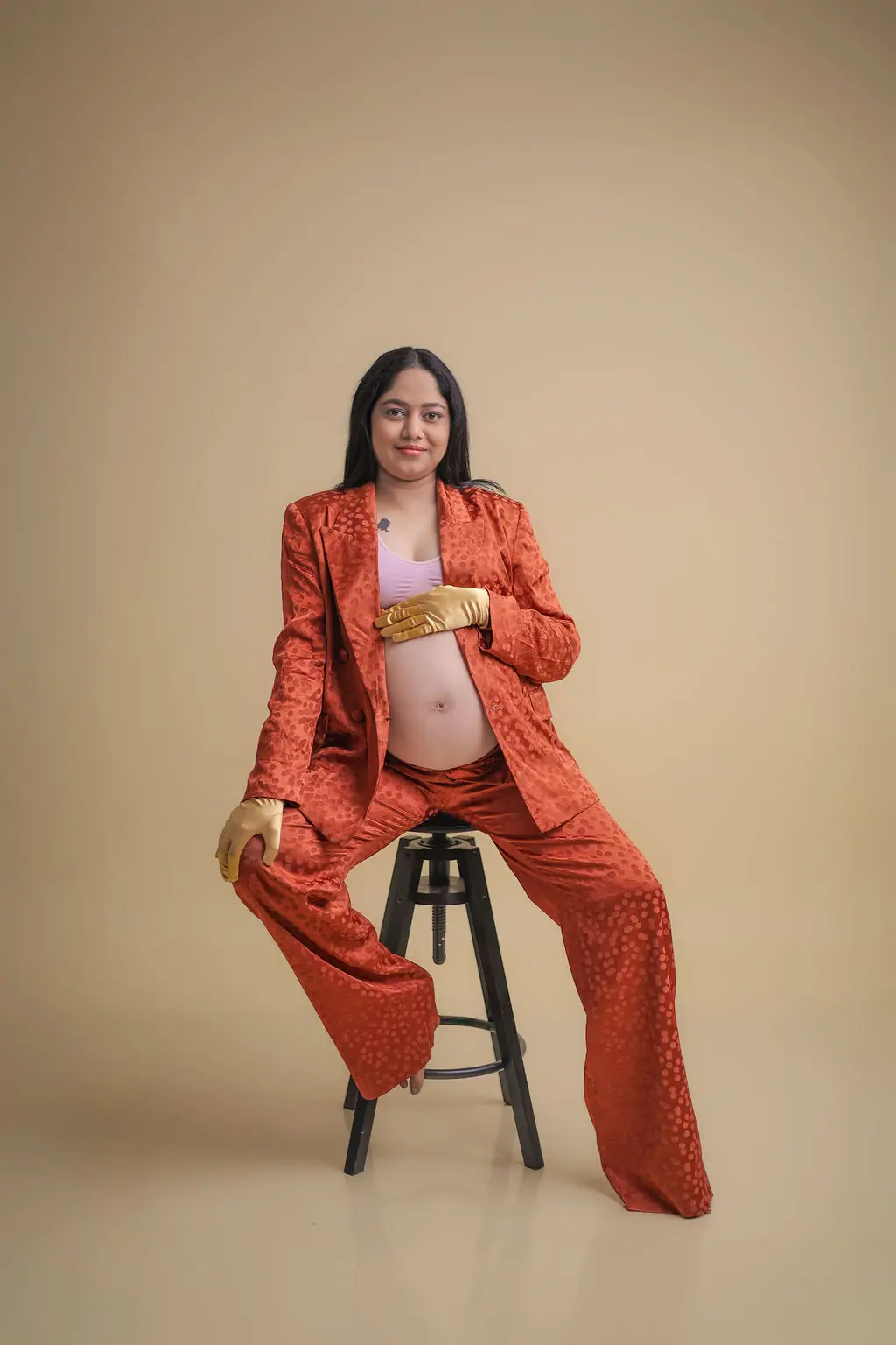 Expectant mother on a black stool in a rust satin suit and gold gloves, beige backdrop, maternity photoshoot at WonderBump Studio, Bengaluru