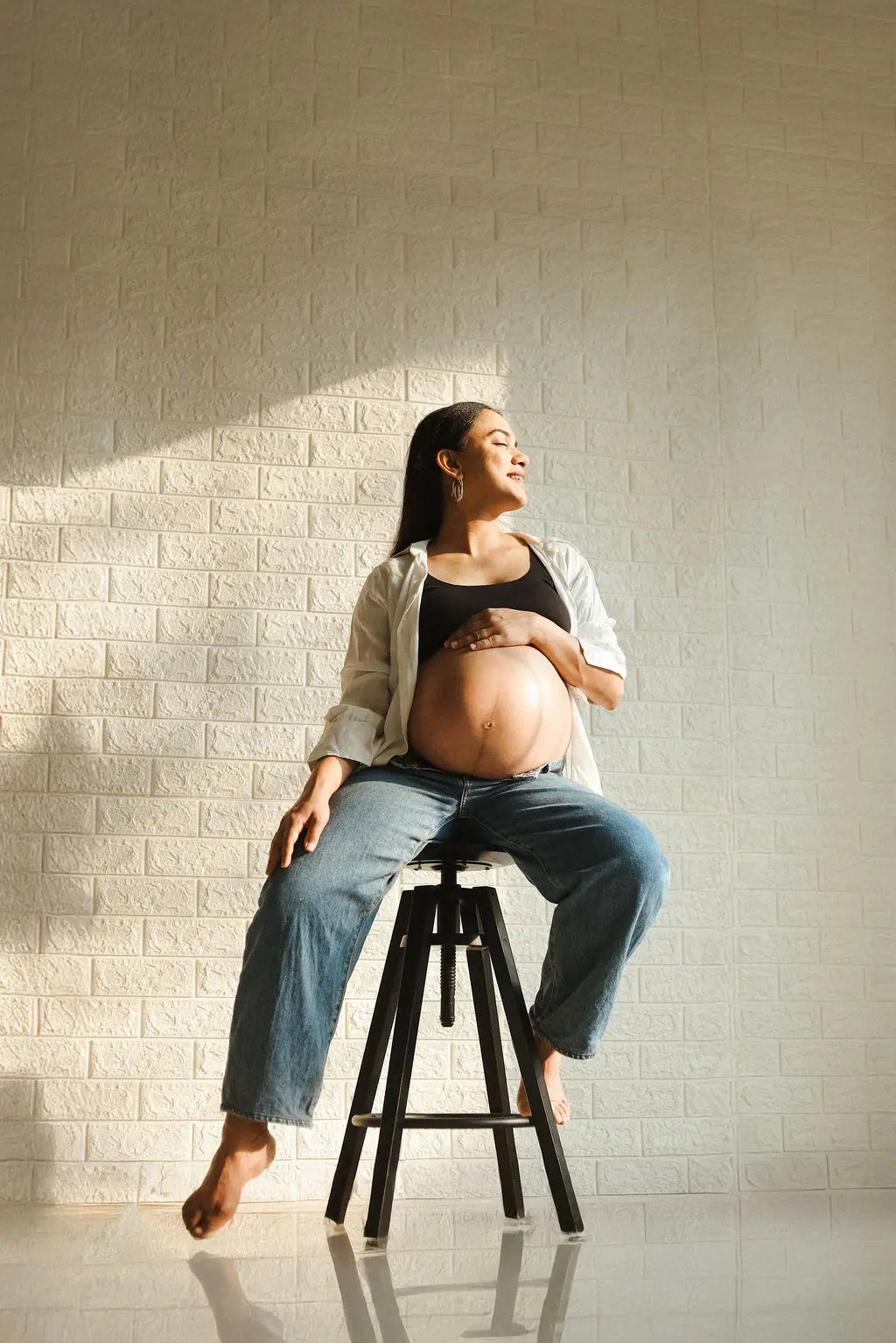 Expectant mother on a black stool in a white shirt and jeans, hand on her bump by a sunlit brick wall, maternity photoshoot at WonderBump Studio, Bengaluru