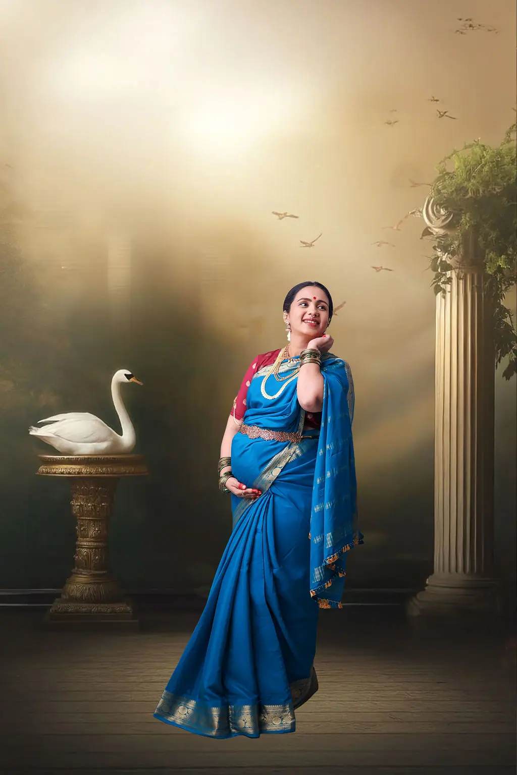 Expectant mother in a blue silk saree beside a swan and column on a printed backdrop, maternity photoshoot at WonderBump Studio, Bengaluru