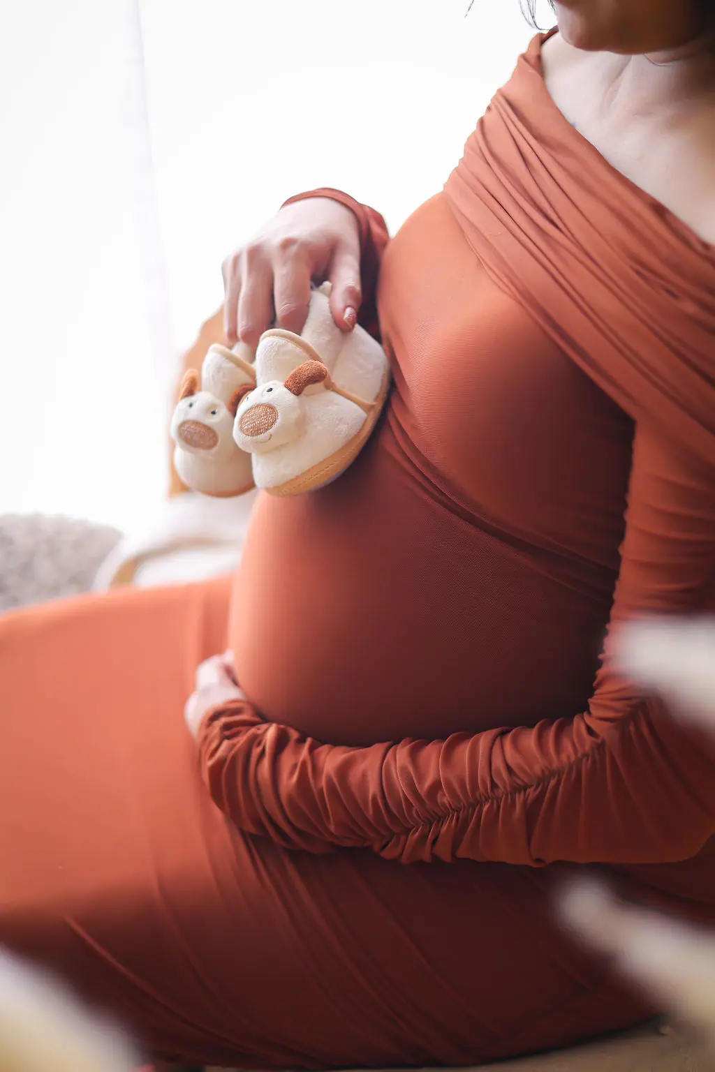 Close-up of an expectant mother in a rust gown holding tiny puppy baby booties against her bump, maternity photoshoot at WonderBump Studio, Bengaluru
