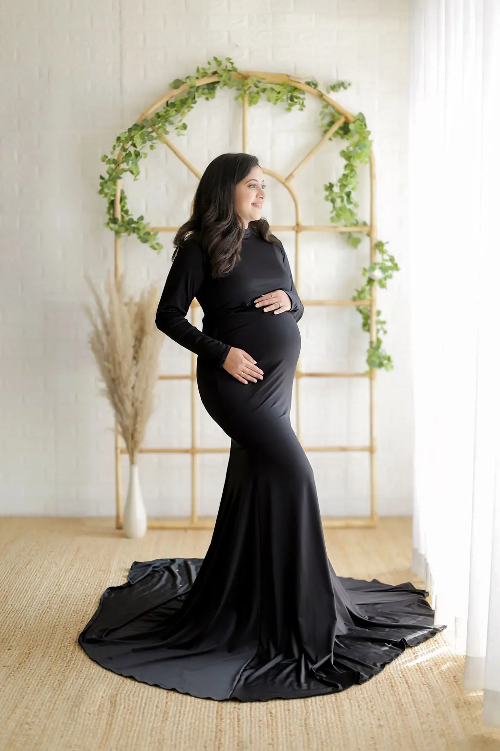 Expectant mother cradling her bump in a black gown with a train by a rattan arch, maternity photoshoot at WonderBump Studio, Bengaluru