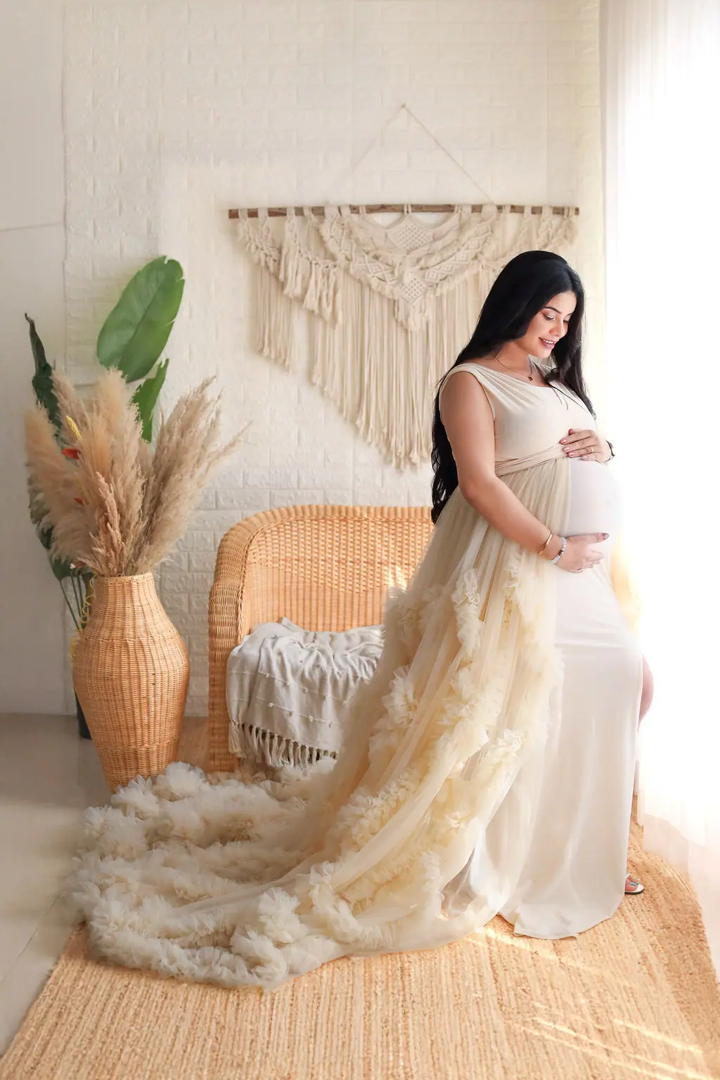 Expectant mother cradling her bump in a cream ruffled gown by a wicker chair, maternity photoshoot at WonderBump Studio, Bengaluru