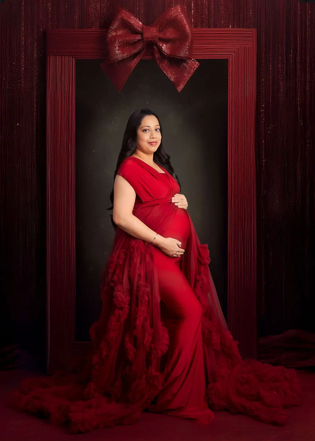 Expectant mother cradling her bump in a red ruffled gown under a giant red bow, maternity photoshoot at WonderBump Studio, Bengaluru