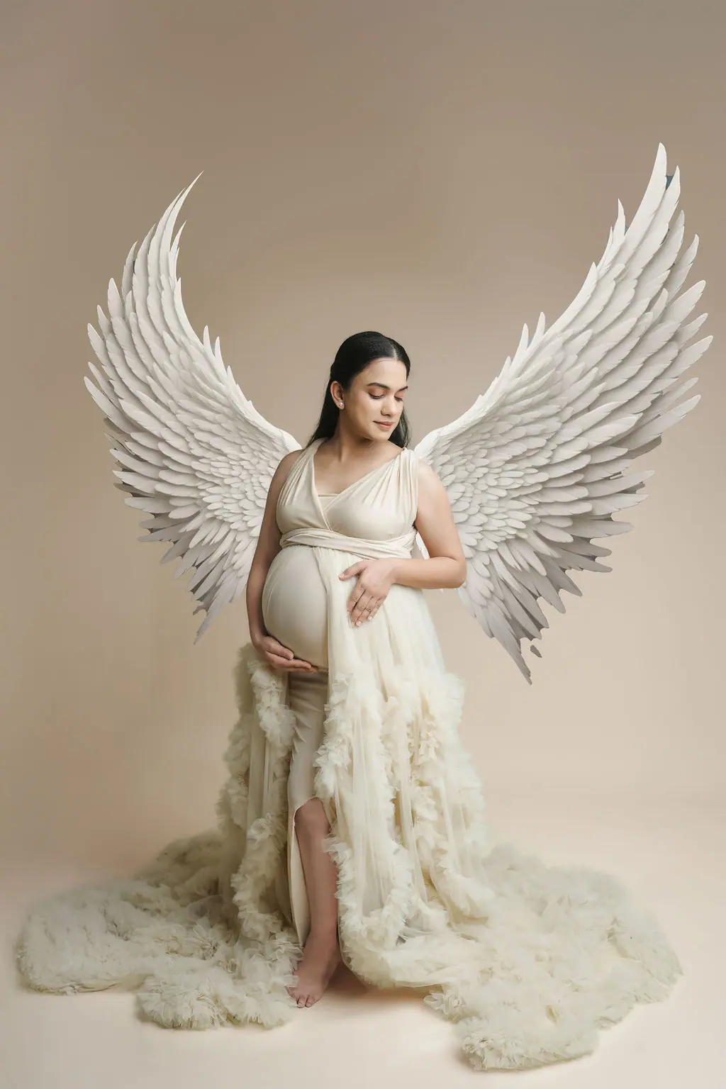 Expectant mother cradling her bump in a cream ruffled tulle gown with large white feathered wings, maternity photoshoot at WonderBump Studio, Bengaluru