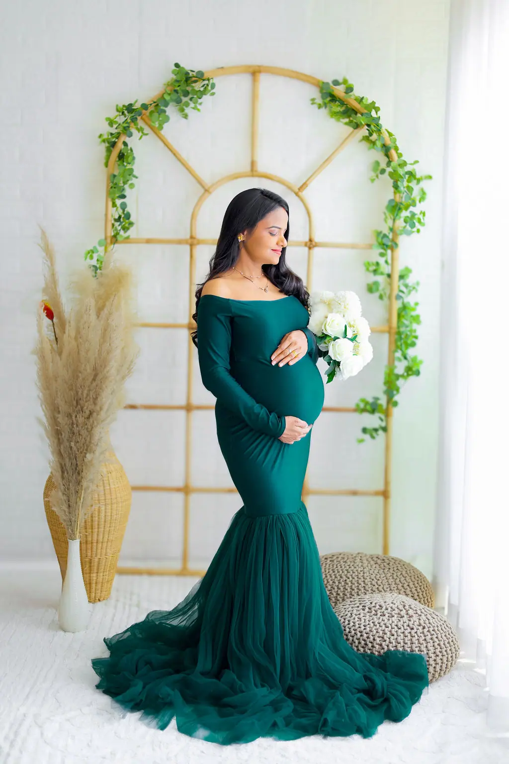 Expectant mother holding white flowers in an emerald tulle gown by a rattan arch, maternity photoshoot at WonderBump Studio, Bengaluru