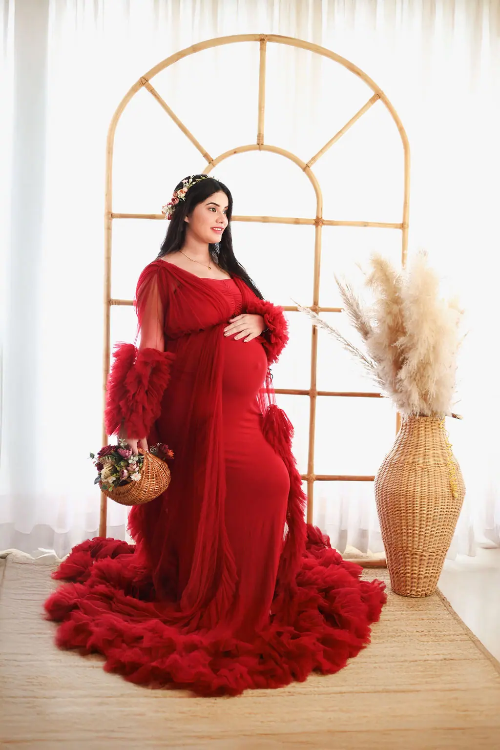 Expectant mother with a flower basket in a red ruffled gown by a rattan arch, maternity photoshoot at WonderBump Studio, Bengaluru