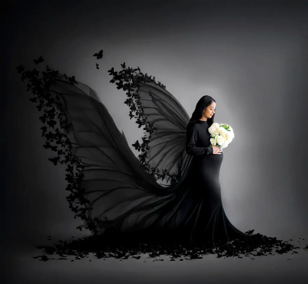 Expectant mother holding white roses in a black gown with a butterfly wing train, maternity photoshoot at WonderBump Studio, Bengaluru