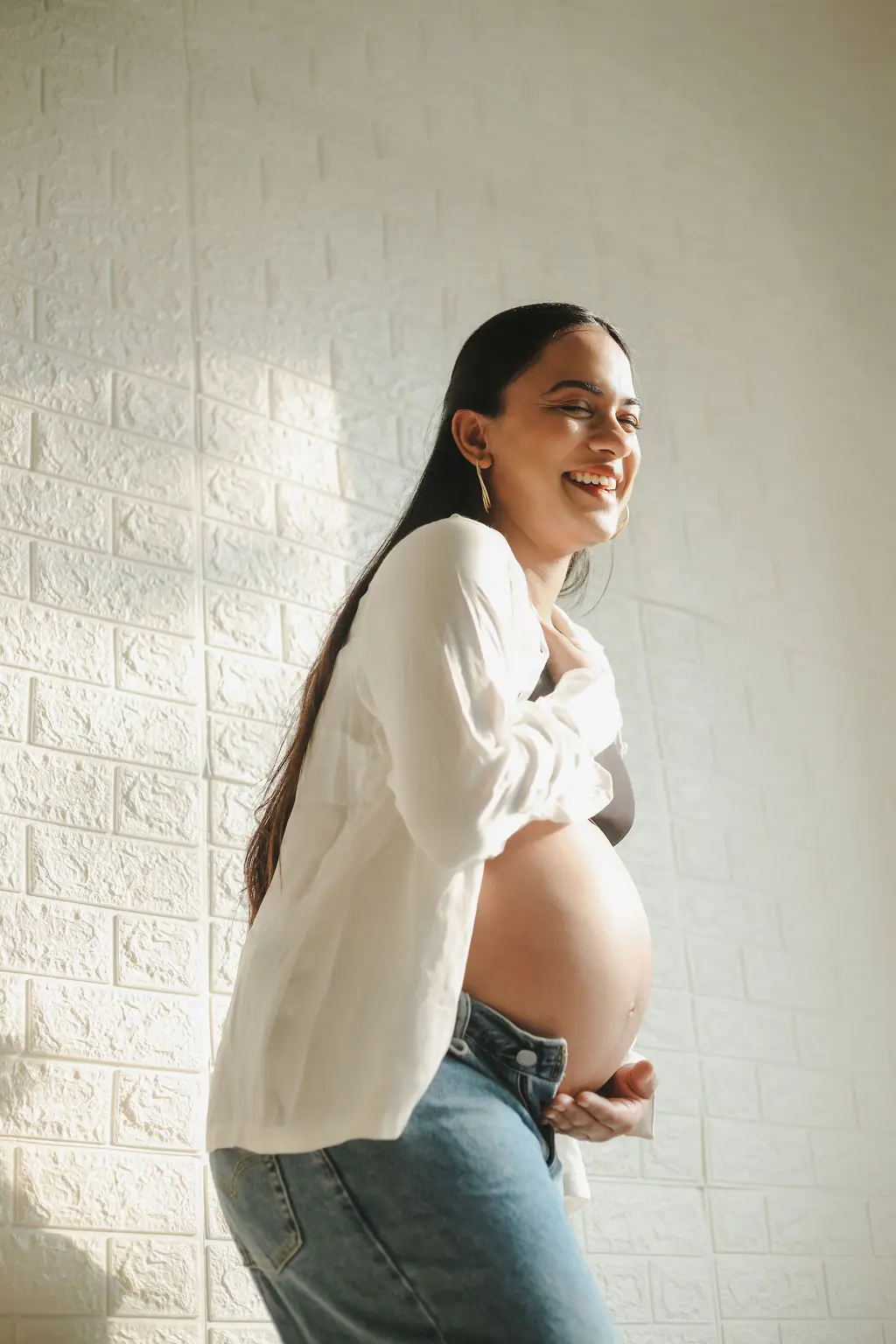 Laughing expectant mother in a white shirt and jeans holding her bump by a sunlit white brick wall, maternity photoshoot at WonderBump Studio, Bengaluru