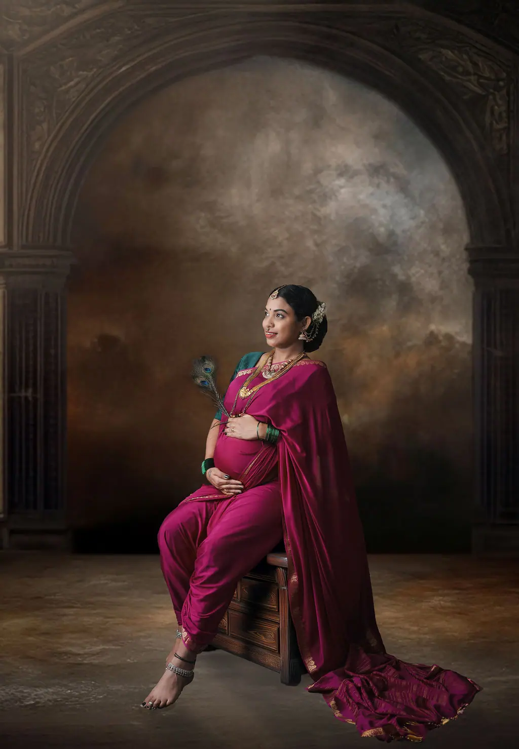 Expectant mother in a magenta nauvari saree seated with a peacock feather, painted arch backdrop, maternity photoshoot at WonderBump Studio, Bengaluru