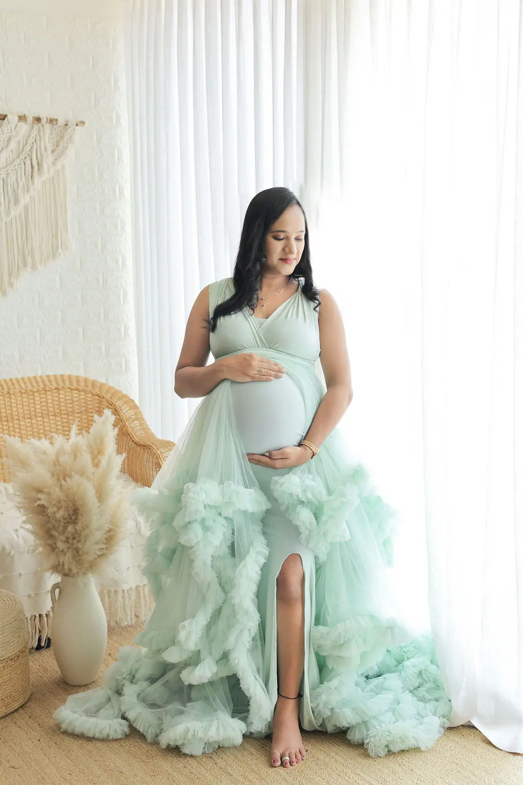 Expectant mother in a mint ruffled tulle gown by sheer curtains, wicker chair and pampas grass, maternity photoshoot at WonderBump Studio, Bengaluru