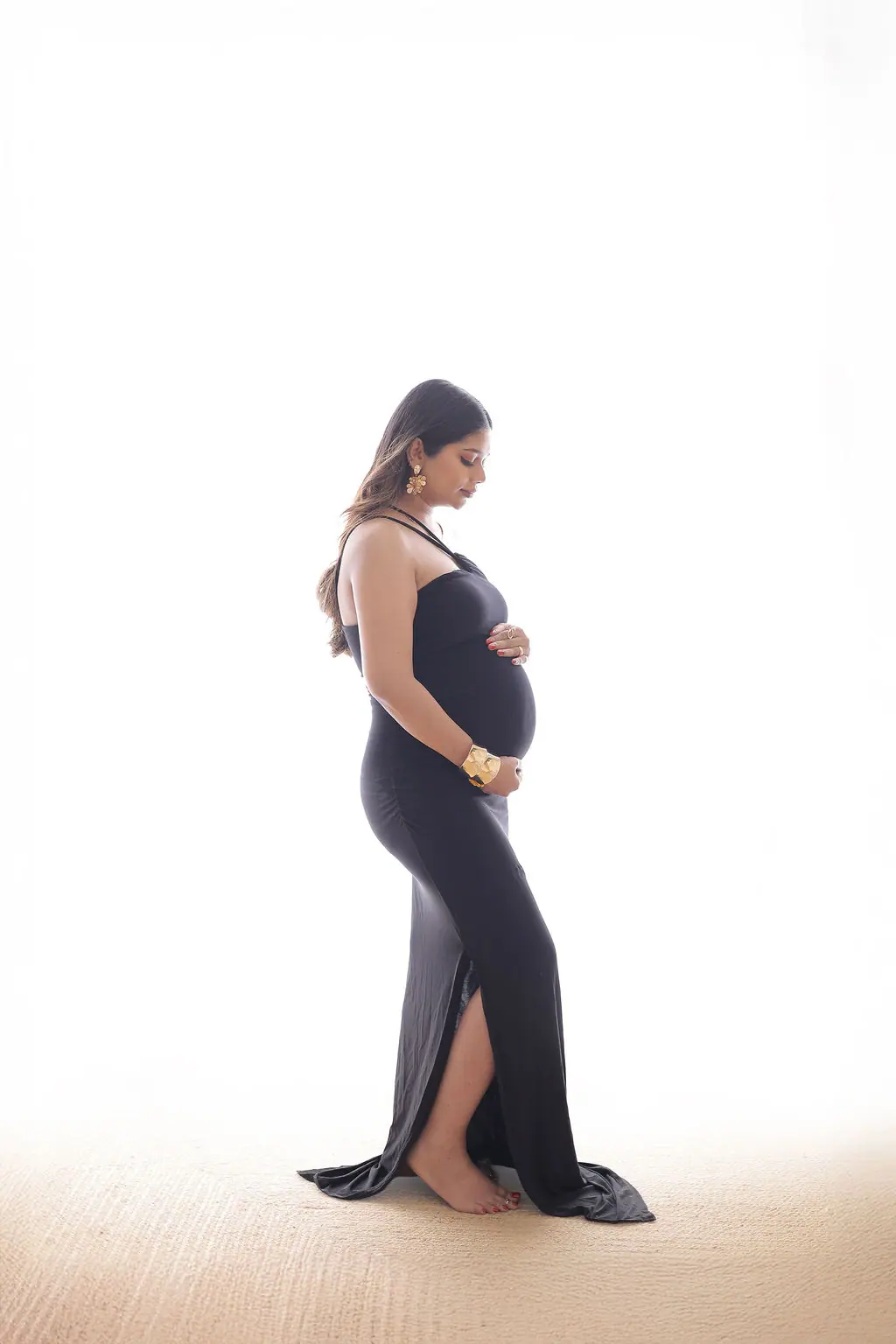 Expectant mother in profile holding her bump in a black one-shoulder gown on a white backdrop, maternity photoshoot at WonderBump Studio, Bengaluru