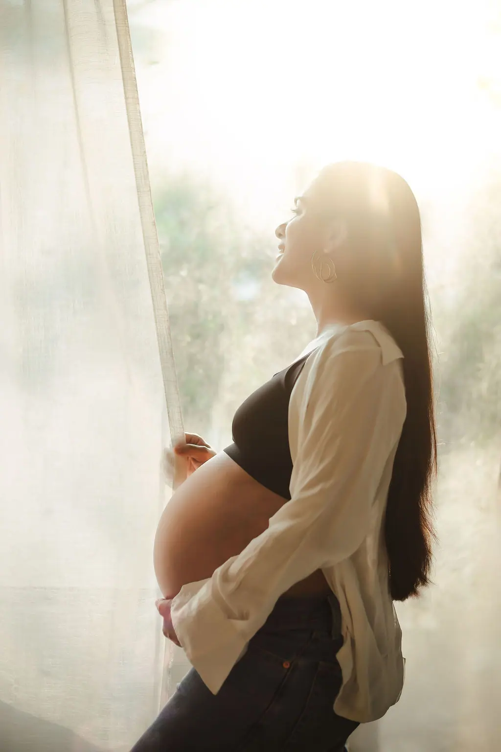 Expectant mother in profile holding a sheer curtain in soft window light, in a white shirt and jeans, maternity photoshoot at WonderBump Studio, Bengaluru
