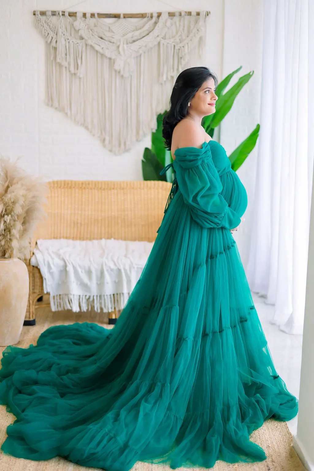 Expectant mother in profile in a teal tiered tulle gown by a window, with a rattan bench and macrame, maternity photoshoot at WonderBump Studio, Bengaluru
