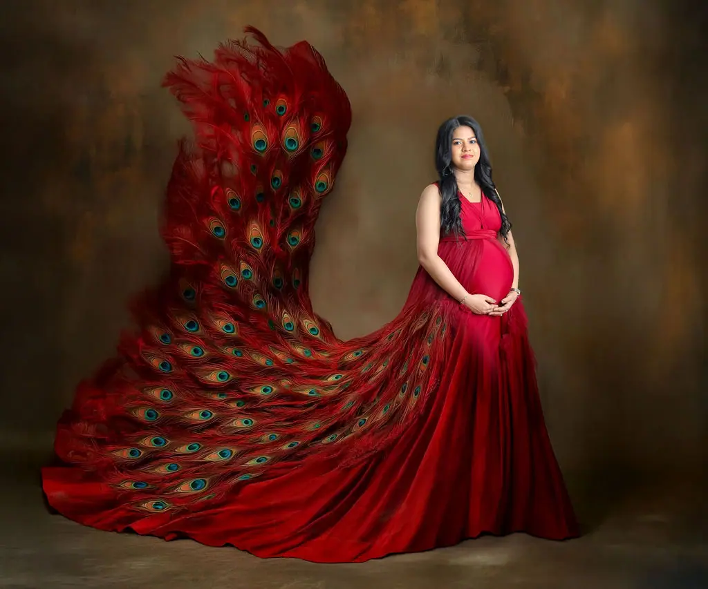 Expectant mother in a red gown with a peacock feather train on a painted backdrop, maternity photoshoot at WonderBump Studio, Bengaluru