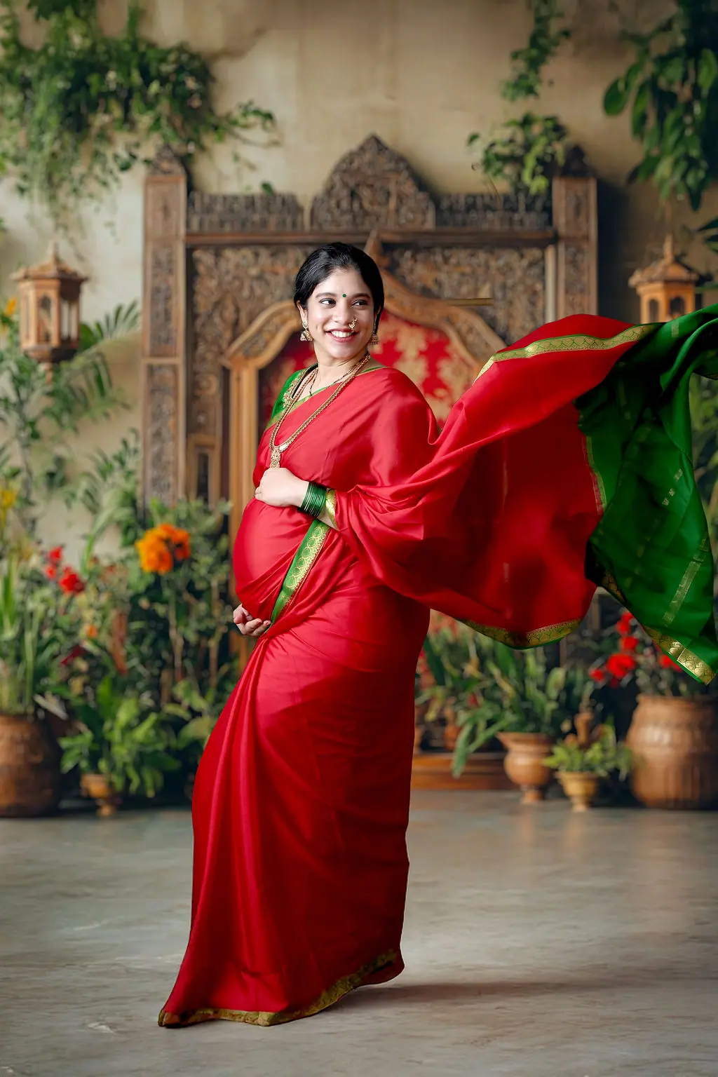 Expectant mother in a red and green silk saree, pallu flying, by a carved wooden arch and plants, maternity photoshoot at WonderBump Studio, Bengaluru