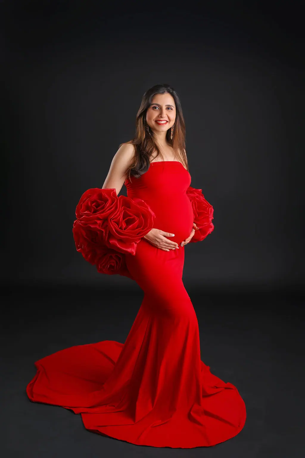 Smiling expectant mother in a red mermaid gown with giant rose sleeves on a black backdrop, maternity photoshoot at WonderBump Studio, Bengaluru