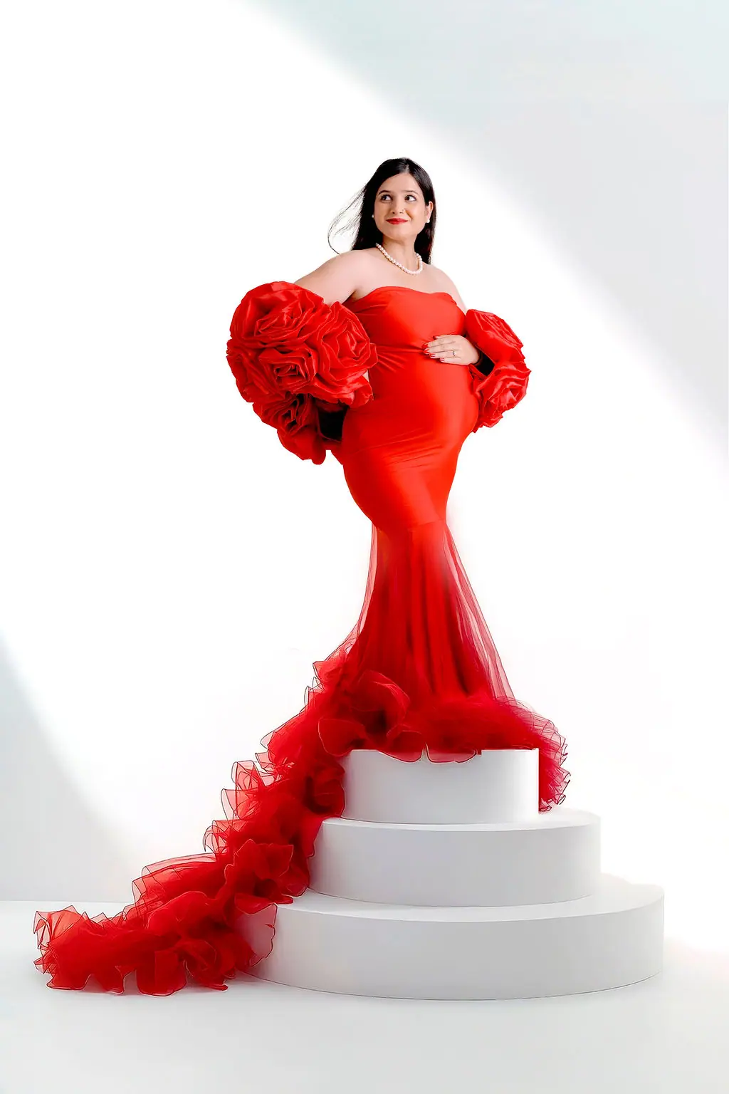 Expectant mother in a red mermaid gown with rose sleeves and a ruffled train on white tiered steps, maternity photoshoot at WonderBump Studio, Bengaluru