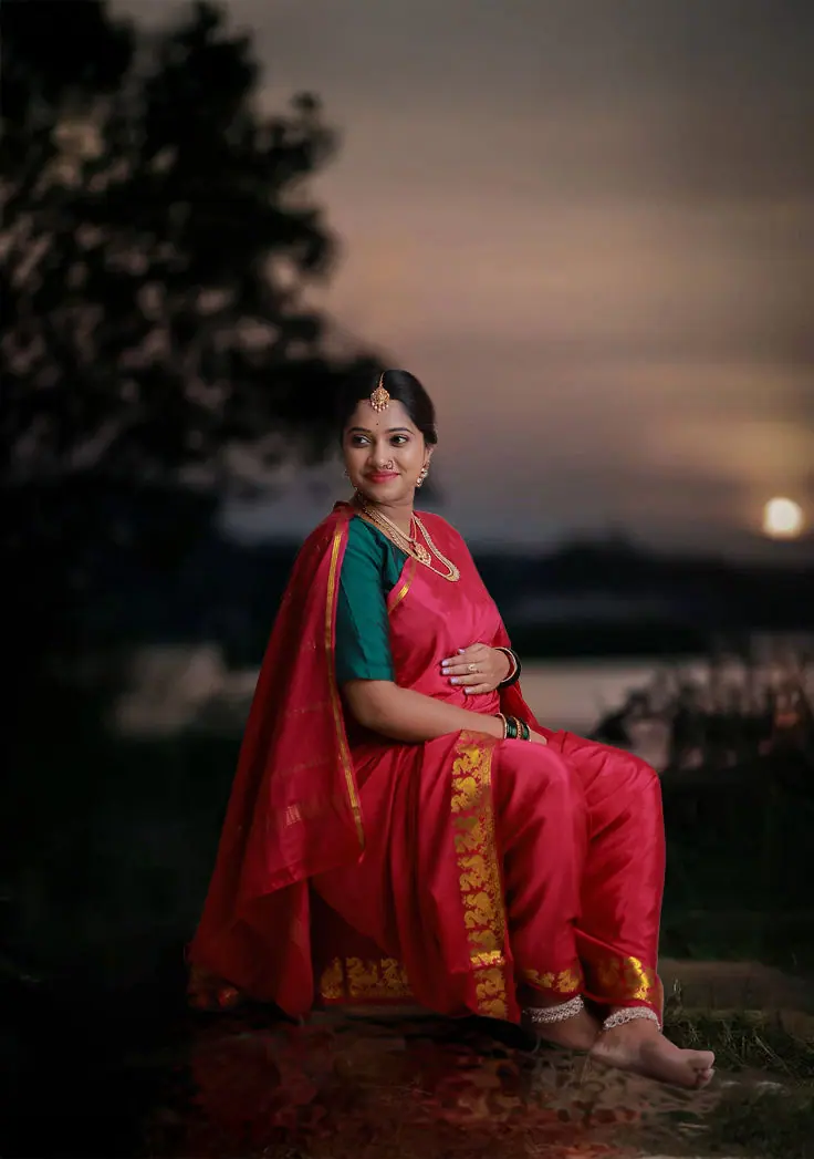 Expectant mother seated in a red silk nauvari saree with a green blouse against a sunset sky, maternity photoshoot by WonderBump Studio, Bengaluru